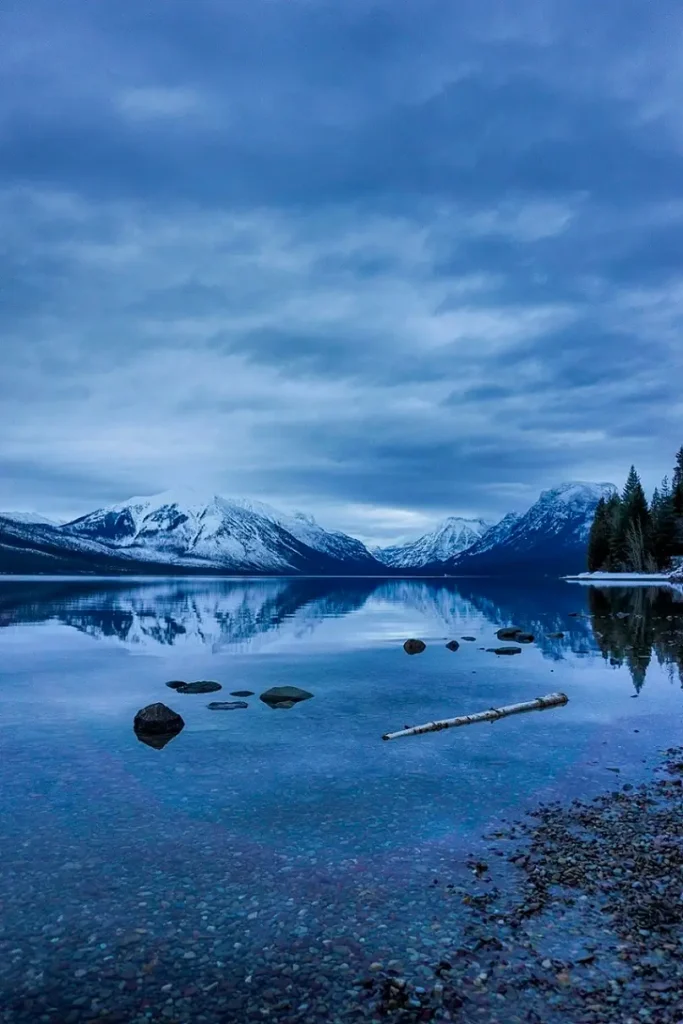 A tranquil scene at Lake McDonald in Glacier National Park, with snow-capped mountains reflected in the still, glassy water. Smooth pebbles are visible beneath the clear lake surface in the foreground, alongside scattered rocks and a driftwood log. The overcast sky adds a moody, cool tone to the landscape, enhancing the serene atmosphere.
