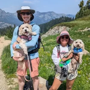 Minerva Samuel Toby Lily Mount Rainier A couple, Samuel and Minerva, pose for a photo on the Pacific Crest Trail at Mount Rainier National Park, each holding a small dog. Samuel, wearing a light blue shirt, pink shorts, and a white wide-brimmed hat, holds Lily, a tan and white dog. Minerva, in a white sweatshirt, pink sunhat, and sunglasses, carries Toby, a yorkie-poo cream-colored dog. They are surrounded by wildflowers and scenic mountain views.