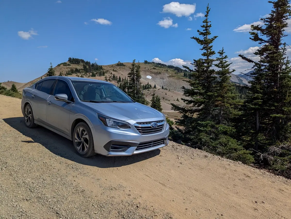 Subaru Legacy Olympic National Park A silver Subaru Legacy is parked on the side of a gravel road of Obstruction Point Road, surrounded by towering pine trees in Olympic National Park. The scene captures the rugged beauty and alpine atmosphere of this high-elevation trail.