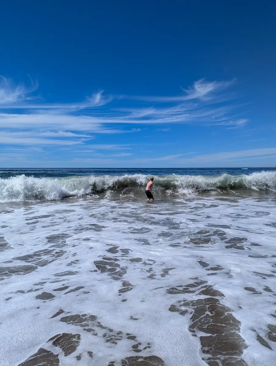Shi Shi Beach Pacific Ocean Sam There A person stands waist-deep in foamy ocean waves at Shi Shi Beach Washington under a clear blue sky streaked with wispy clouds, enjoying the surf on a sunny day.