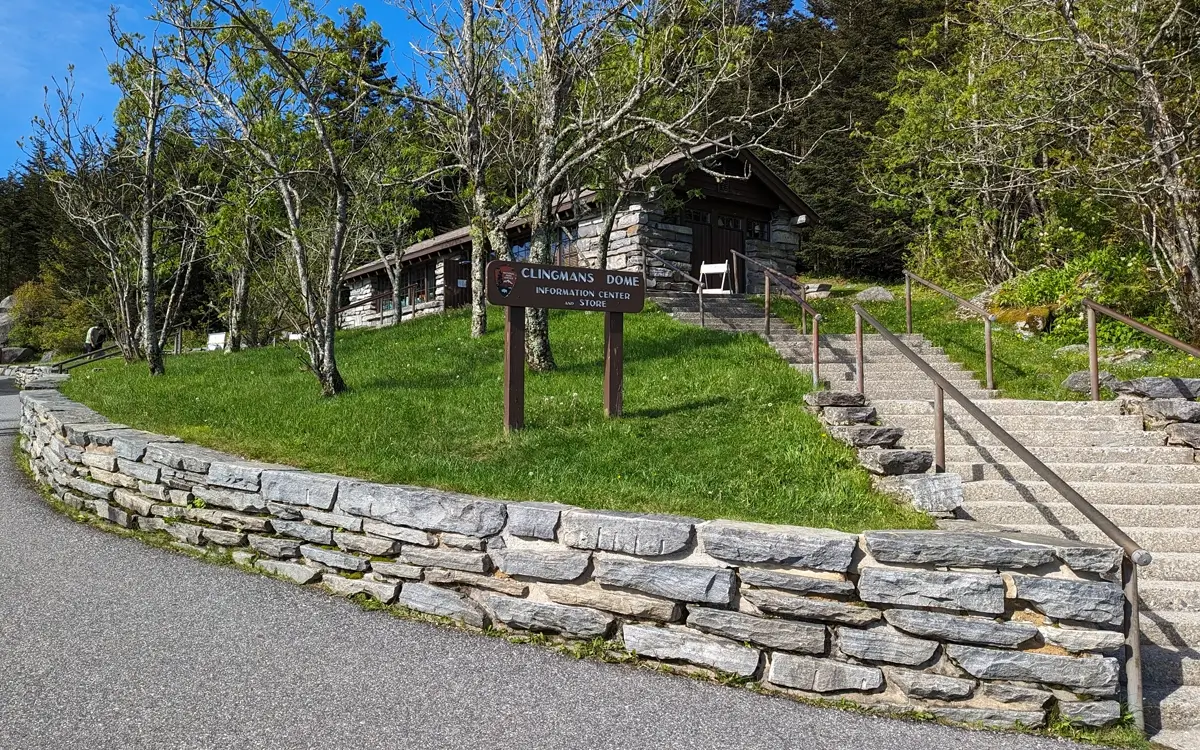 Clingmans Dome Information Center A rustic log-and-stone visitor center marked with a brown sign reading “Clingmans Dome Information Center and Store,” surrounded by greenery, trees, and a stone-lined pathway with steps leading up.