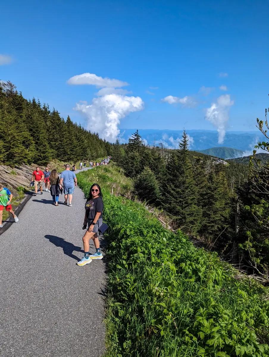 Clingmans Dome Trail Hikers walk along the paved Clingmans Dome trail lined with lush green vegetation and dense evergreen trees, as distant clouds rise dramatically against the backdrop of blue mountains.
