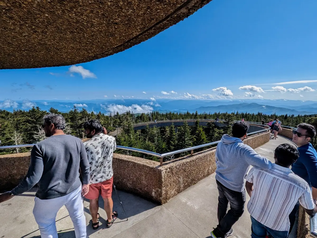 Kuwohi Observation Area Visitors stand along the curved concrete ramp leading to the Clingmans Dome observation tower, enjoying panoramic views of the Blue Ridge Mountains under a bright blue sky with scattered clouds.