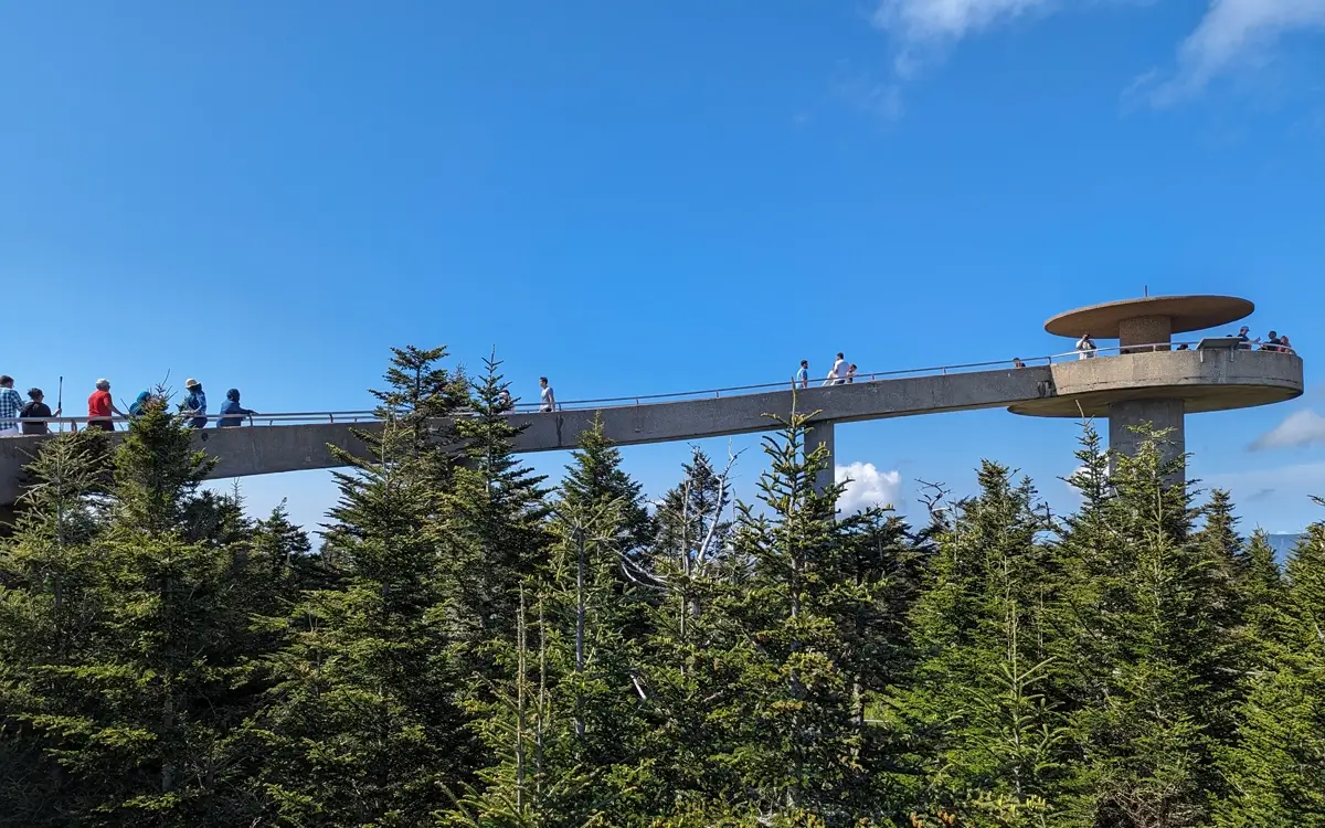 Side view of Kuwohi Observation Tower with its circular platform perched atop a tall concrete pillar, connected to a ramp filled with visitors walking above a lush forest of spruce-fir trees on a clear sunny day.