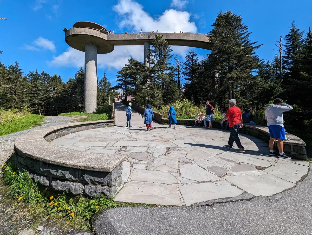 Kuwohi Observation Tower Great Smoky Mountains Ground-level view of the base of Kuwohi Observation Tower showing a curved stone plaza with benches, visitors resting or walking, and the ramp ascending through the trees toward the iconic round observation platform.