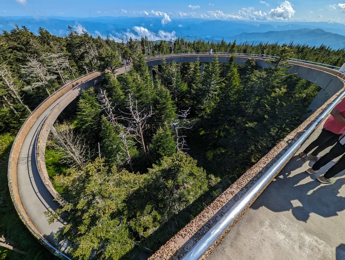 Kuwohi Tower Spiral A sweeping aerial view of the curved, spiral walkway of Clingmans Dome Observation Tower, surrounded by dense evergreen forest with hazy blue mountains layered in the distance under a bright sky.
