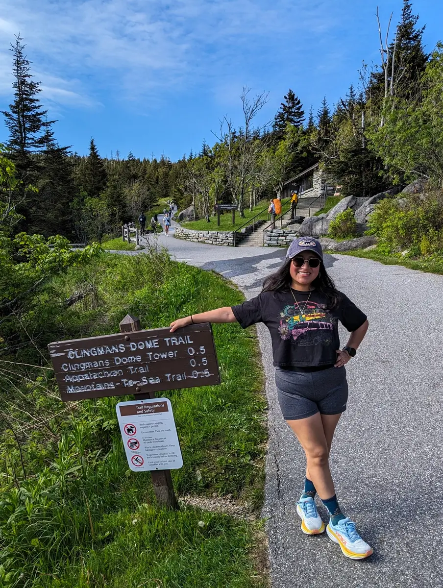 Minerva Standing On Clingmans Dome Trail A woman in sunglasses and a graphic t-shirt poses with one arm resting on a wooden sign marking the Clingmans Dome Trail, surrounded by lush greenery and hikers in the distance; the sign points to Clingmans Dome Tower, the Appalachian Trail, and the Mountains-to-Sea Trail.