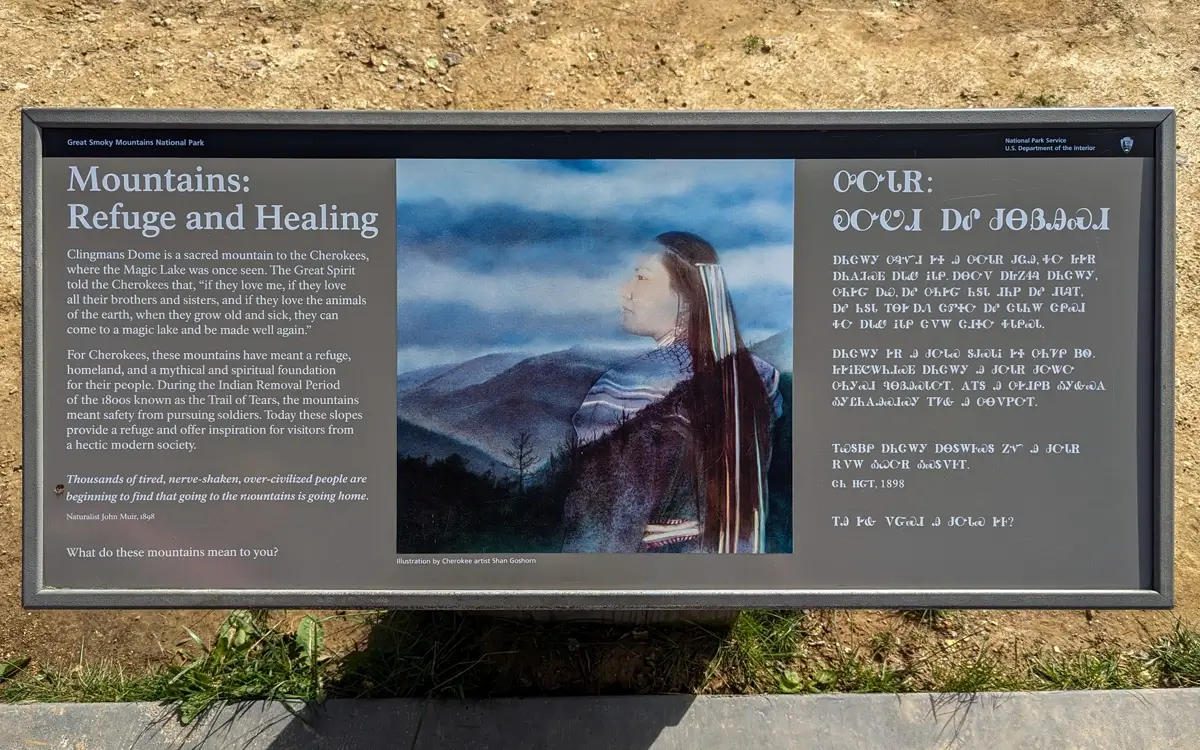 Mountains Refuge And Healing An interpretive sign titled “Mountains: Refuge and Healing” describes the cultural and spiritual importance of Clingmans Dome to the Cherokee people. The sign includes a quote from John Muir, a painting of a Cherokee figure gazing over the mountains, and text in both English and the Cherokee syllabary. This sign is located along the Kuwohi Observation Tower Trail.