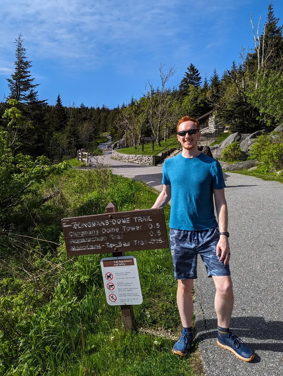 Sam Standing On Clingmans Dome Trail A smiling man in a blue shirt and shorts stands next to a wooden trail sign at Clingmans Dome, with a winding paved path and dense forest in the background. The sign points along the Kuwohi Observation Tower trail to destinations including Clingmans Dome Tower, the Appalachian Trail, and the Forney Ridge Trail.