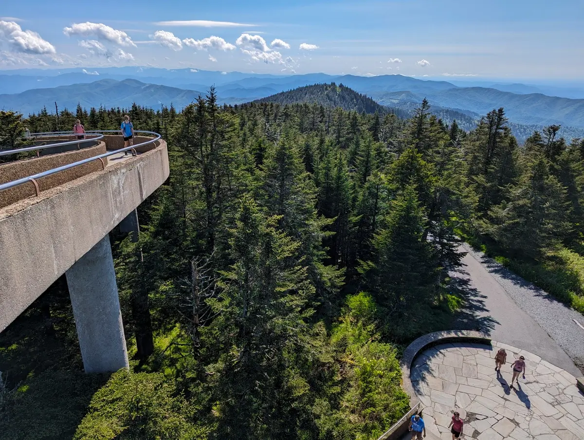 View From Kuwohi Tower A scenic view from Clingmans Dome Observation Tower shows visitors walking along the concrete ramp and stone plaza below, surrounded by dense evergreen forest, with layers of blue mountain ridges stretching into the horizon under a bright, cloud-speckled sky.