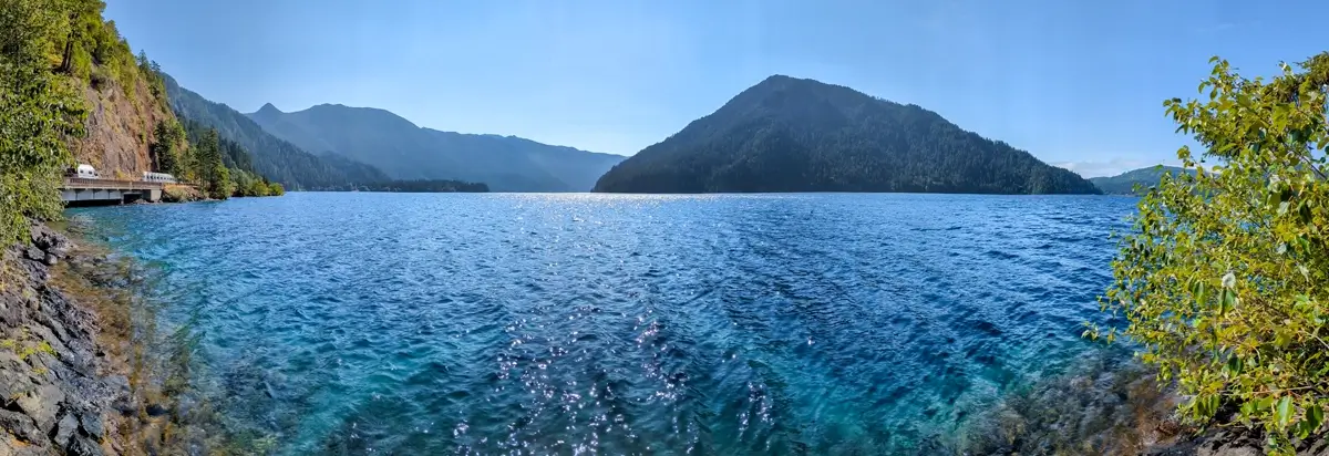 Lake Crescent Panorama A scenic panorama of Lake Crescent Washington from Highway 101. The clear blue water stands prominently in the foreground surrounded by green foliage with mountains cascading into the distance under a blue sky.