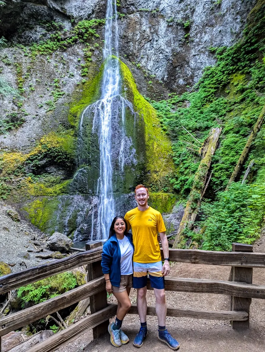 Minerva And Sam Standing In Front Of Marymere Falls Washington Two hikers, one wearing a yellow shirt with red hair and the other in a blue sweatshirt, stand in front of Marymere Falls, a 90-foot tall, narrow waterfall in Olympic National Park. The area is surrounded by lush forest and fallen logs. A wooden railing is behind the hikers.