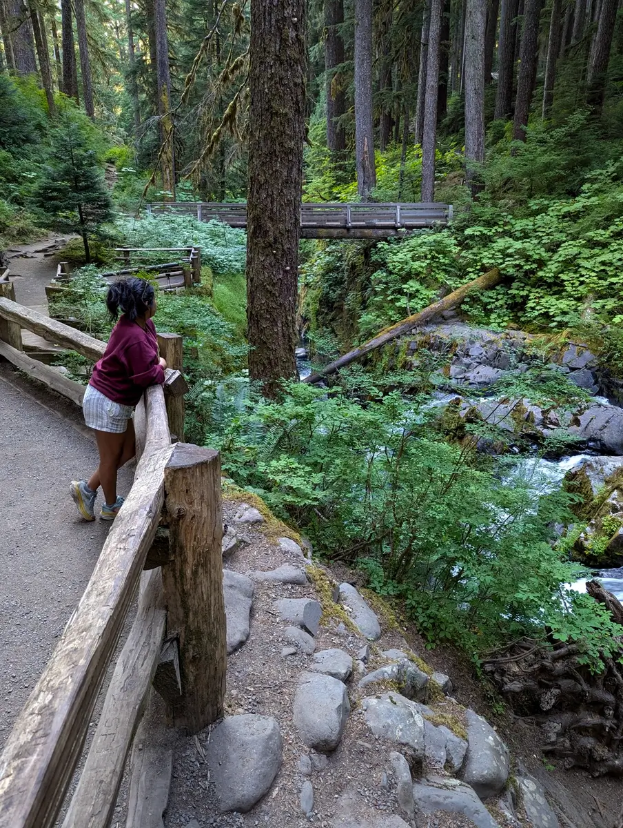 A woman in a burgundy sweater and plaid shorts leans against a rustic wooden railing, gazing at a forested ravine with Sol Duc Falls and wooden footbridge in the distance. Lush green foliage and tall trees surround the tranquil trail.