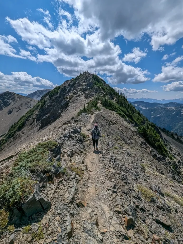 A solo hiker walks along a narrow, rocky ridgeline trail leading toward a rugged mountain peak, with dramatic clouds casting shadows over the surrounding mountain ranges of Olympic National Park.
