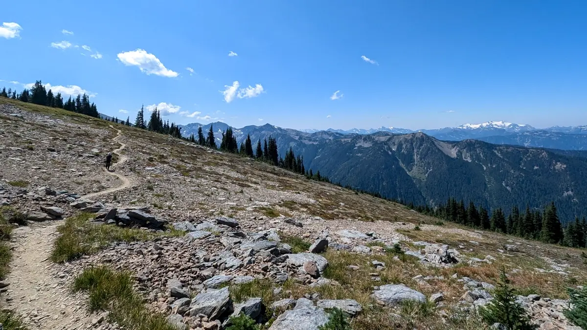 Grand Valley Trail A dirt hiking trail winds through an alpine meadow scattered with rocks and evergreens, with jagged mountain ridges and snowy peaks stretching into the distance under a bright blue sky.