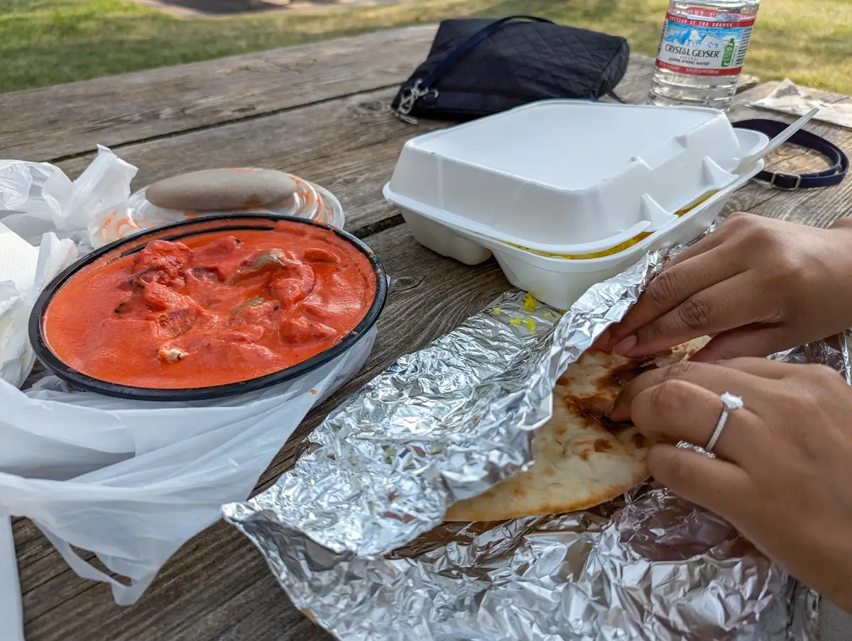 Maryhill State Park Picnic Setup Takeout Indian food set on a rustic picnic table at Maryhill State Park, featuring a tray of vibrant orange chicken tikka masala, foil-wrapped naan being opened by a woman wearing a ring, and a styrofoam container of yellow rice in the background.