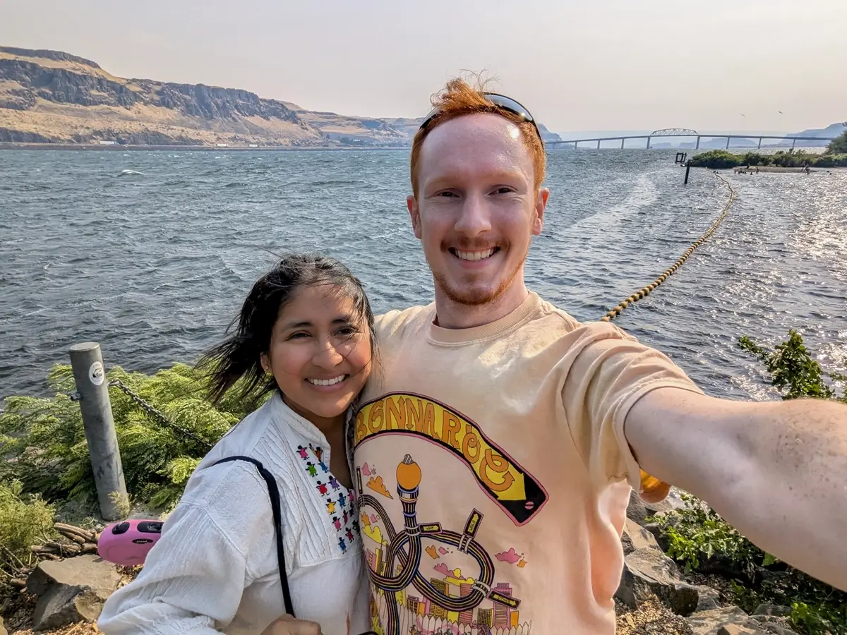 Minerva And Sam At Maryhill State Park A smiling couple takes a selfie by the Columbia River at Maryhill State Park, with wind blowing through their hair and the scenic Sam Hill Memorial Bridge, rugged hills, and rippling water in the background.