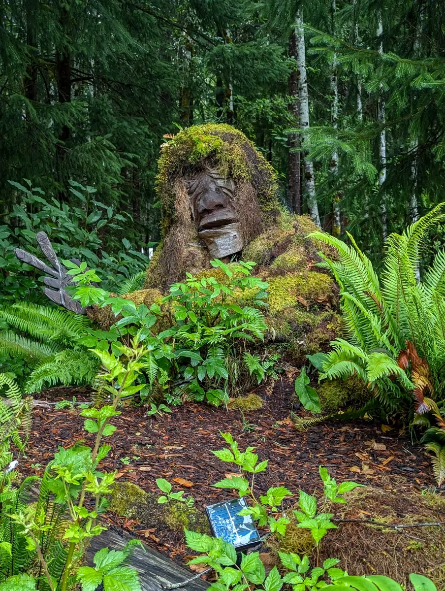 Mossquatch Creature A large Bigfoot statue partially camouflaged with moss and natural materials, emerging from dense ferns and greenery in the forest, blending into its Olympic Peninsula surroundings.
