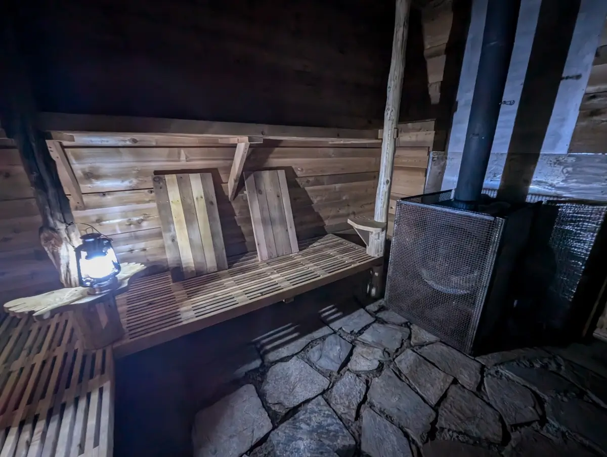 Mossquatch Sauna A cozy wooden sauna with slatted bench seating, a glowing lantern on a carved wooden shelf, a wood-burning stove in the corner, and a stone-tiled floor, creating a warm, off-grid ambiance.