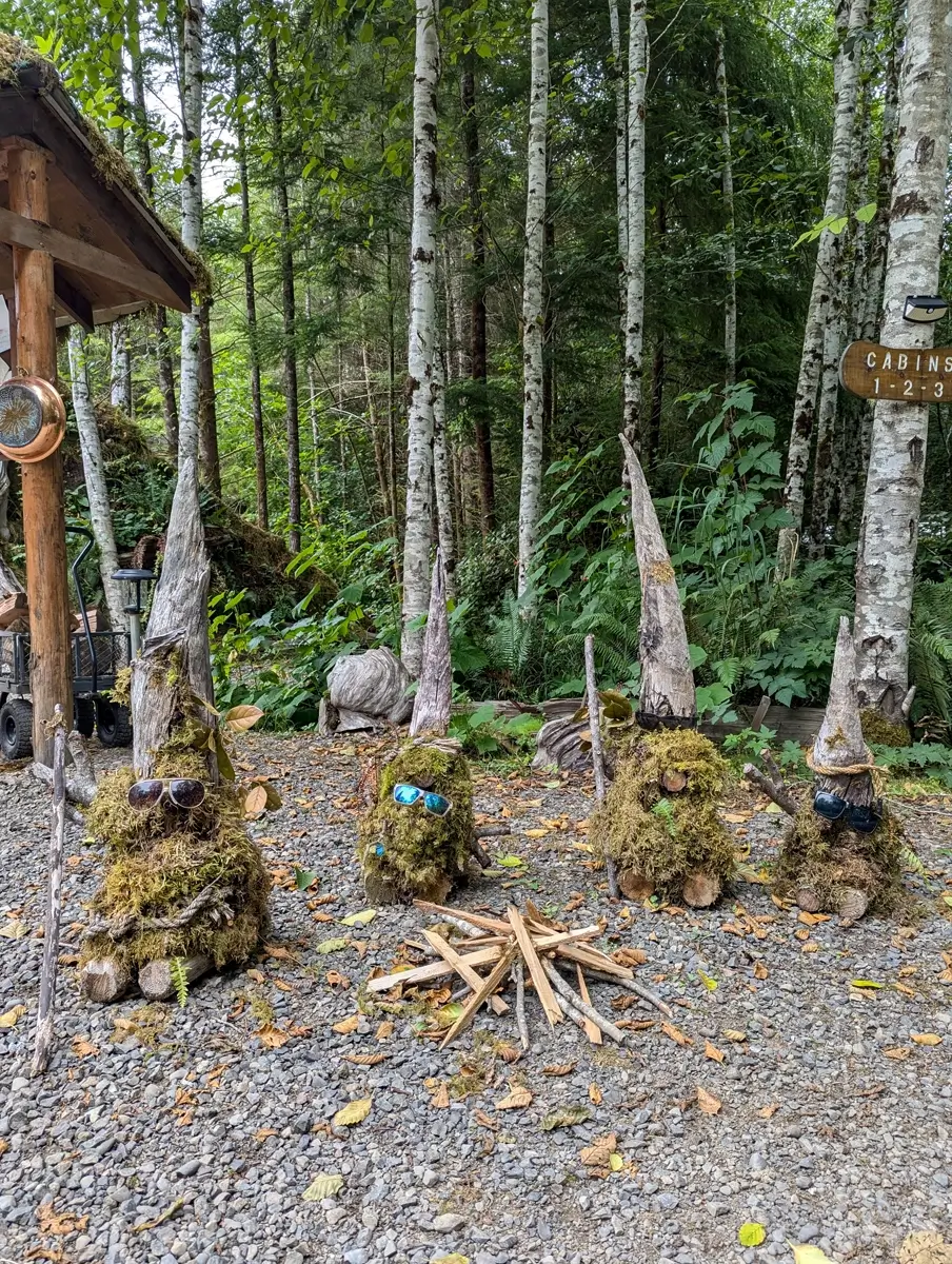 Mossy Gnomes At Mossquatch Whimsical moss gnomes with sunglasses gather around a campfire setup on a gravel path, marking the entrance to cabins at Mossquatch Resort, a unique glamping and cabin retreat near La Push, Washington.