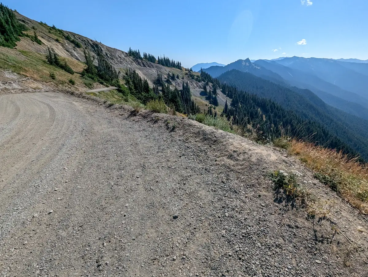 Obstruction Point Road From Hurricane Ridge A winding gravel road curves along a steep mountain slope lined with evergreens, overlooking layers of ridges fading into the hazy blue horizon in Olympic National Park. Obstruction Point Road makes its way towards the trailhead.