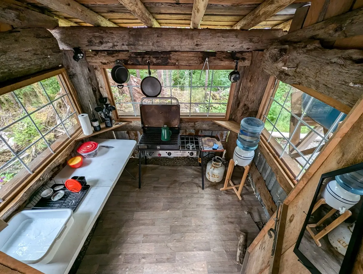 Outdoor Kitchen At Mossquatch A compact rustic kitchen with a propane stove, enamel coffee pot, hanging pans, a water jug station, and basic cooking supplies, surrounded by large windows offering views of lush forest greenery.