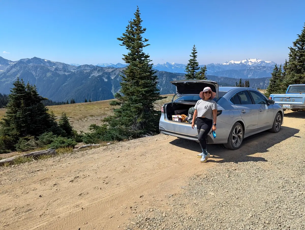 Parked At The Obstruction Point Trailhead A hiker in a sunhat leans casually against the open trunk of a silver car, parked on a dirt pull-off with mountain peaks and alpine forest in the background near the Obstruction Point Trailhead.