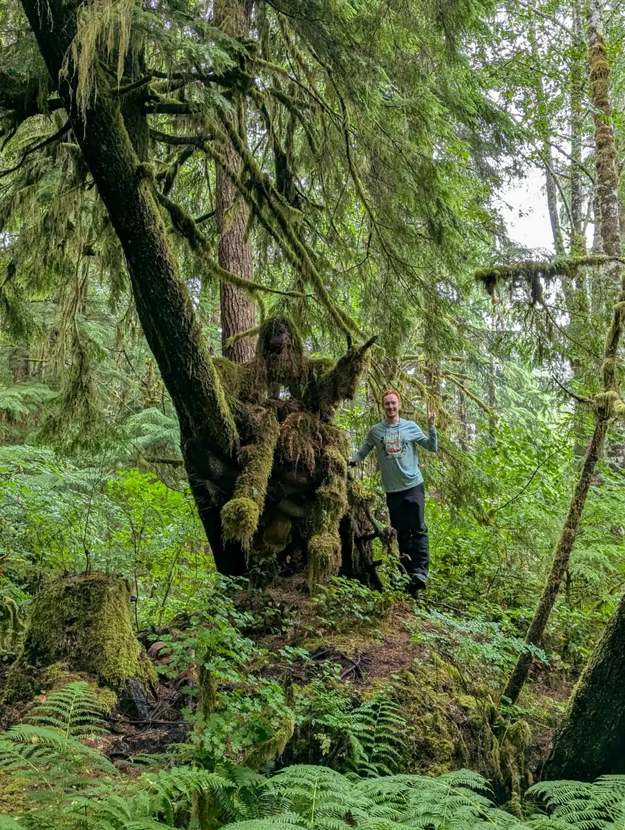 Pointing Mossquatch Creature A visitor poses next to a giant, moss-covered "Mossquatch" creature sculpture in the dense, enchanting forest of Mossquatch Resort, a nature destination on the Olympic Peninsula.