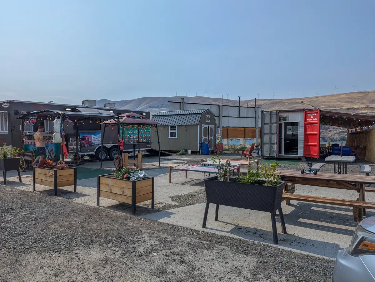 Punjabi Dhaba Food Truck A wide view of the food truck area in Biggs Junction shows a person ordering at the Sizzling Tandoori Hut trailer, surrounded by picnic tables, potted plants, and a converted red shipping container labeled “Welcome to Punjabi Dhaba,” with golden hills and wind turbines in the background.