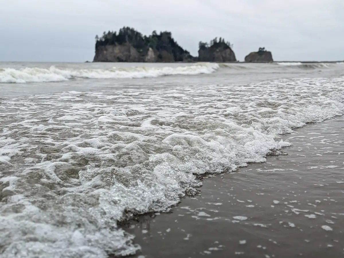 Cresting Waves La Push Close-up of white, foamy waves cresting and rolling onto the sandy First Beach in La Push, Washington, with sea stacks visible in the distance.