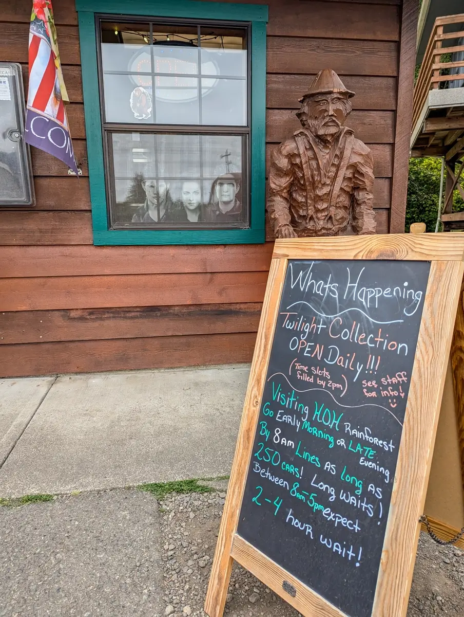 Forks Chamber Of Commerce Wooden storefront with a Twilight-themed poster in the window, a carved lumberjack statue, and a chalkboard sign announcing Twilight Collection hours and visitor tips for the Hoh Rainforest.