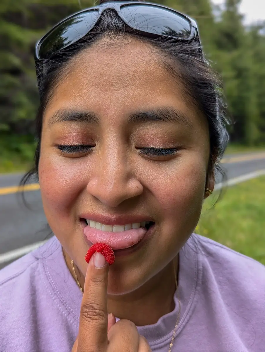 Minerva Eating A Thimbleberry Close-up of a smiling woman with eyes closed, wearing sunglasses on her head, touching her tongue to a fresh raspberry balanced on her fingertip, with a forested roadside in the background.