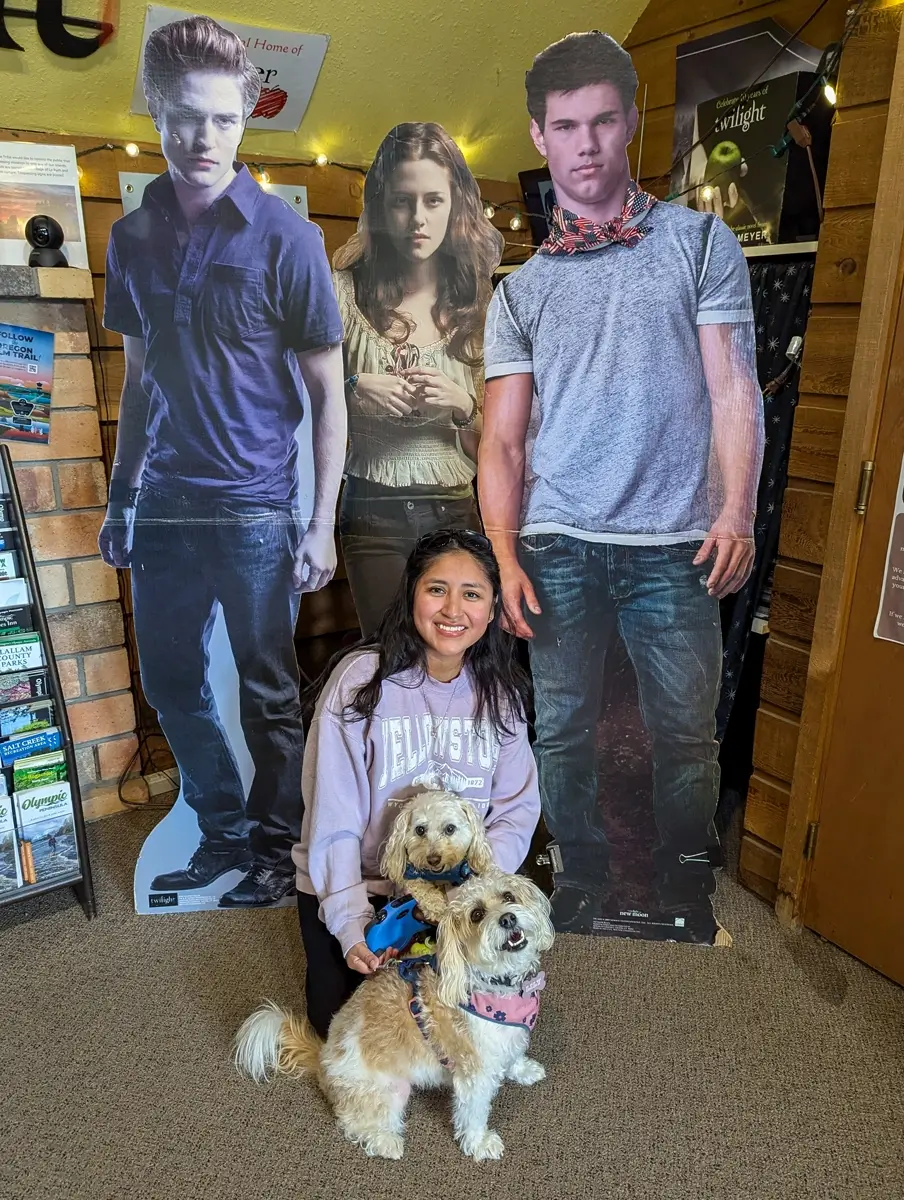 Minerva Toby And Lily With Twilight Cardboard An image of a smiling woman with two small dogs, one with a blue harness and one with a pink floral harness, kneeling in front of three life-sized cardboard cutouts of the Twilight characters Edward, Bella, and Jacob.
