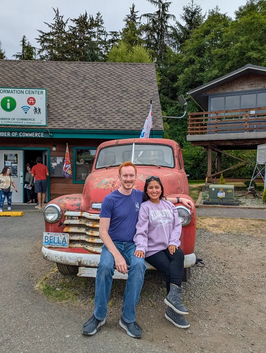 Minerva And Sam With Bellas Twilight Book Truck Man and woman sitting together on the bumper of a weathered red vintage truck with a “BELLA” license plate outside the Forks Chamber of Commerce building. Bella's book truck from Twilight.