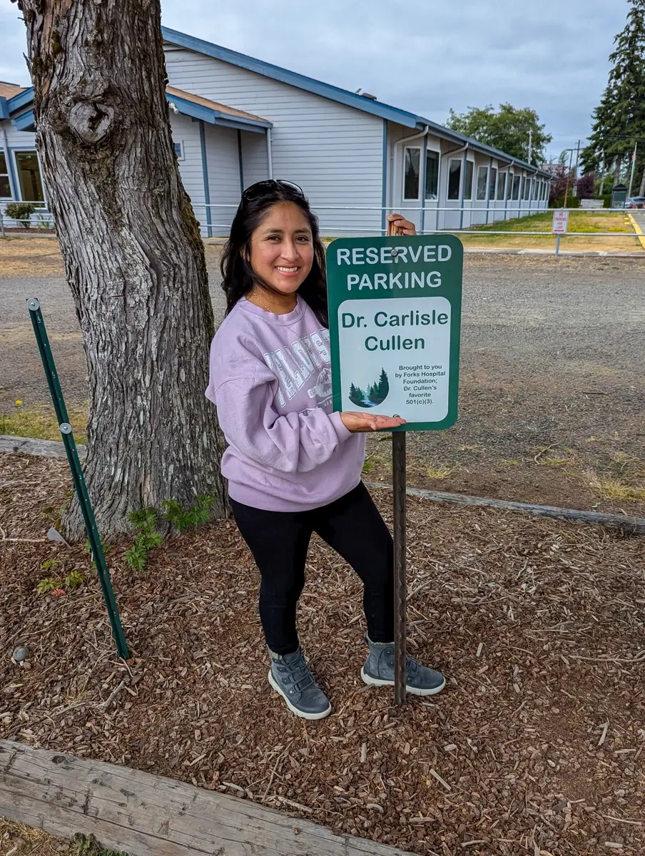 Minerva At Dr. Cullens Parking Forks WA Woman smiling and holding a green reserved parking sign for “Dr. Carlisle Cullen” in front of a light blue building, referencing the Twilight book series.