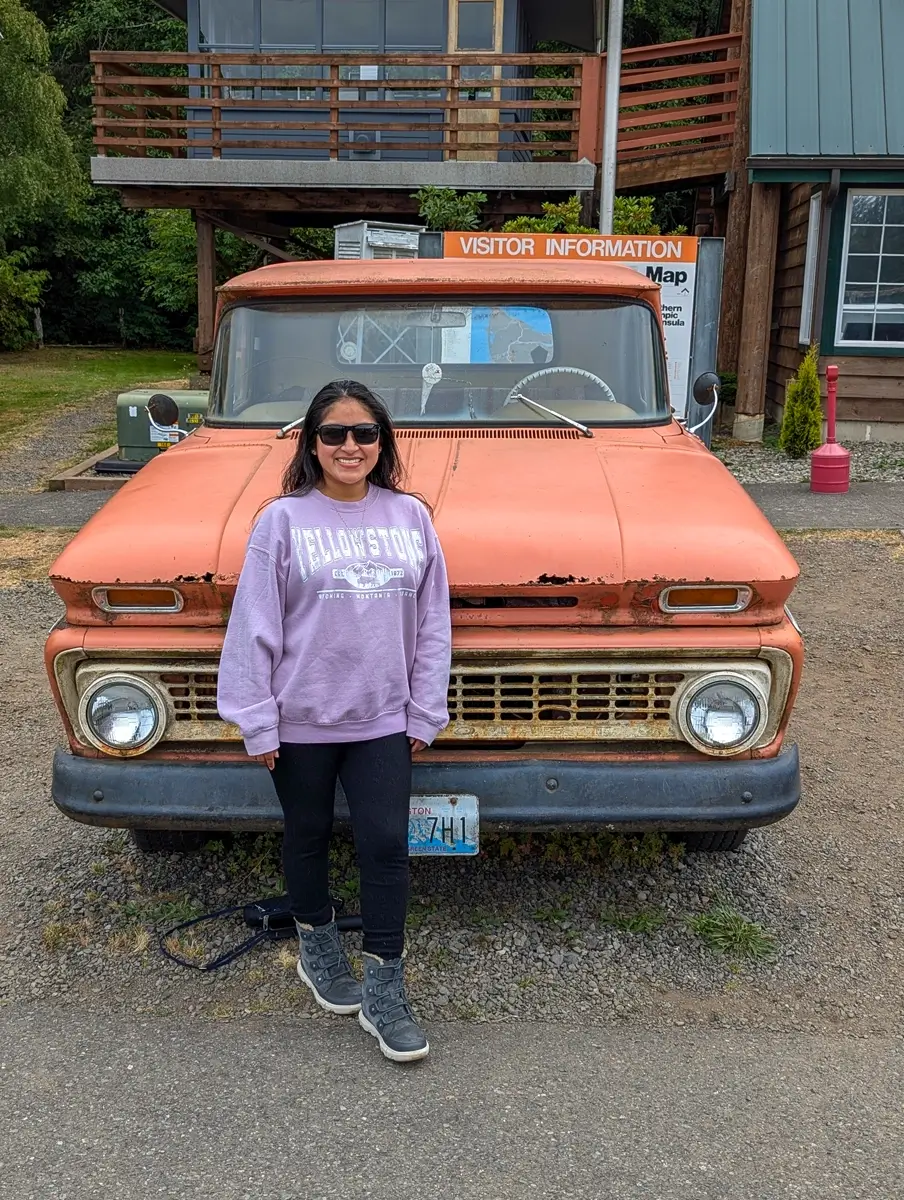 Minerva With Bellas Twilight Movie Truck A woman wearing sunglasses and a purple "Yellowstone" sweatshirt stands in front of an old, orange pickup truck, similar to the one driven by Bella Swan in the movie "Twilight."
