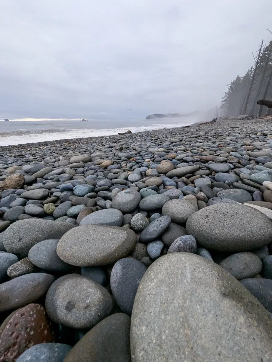 A close-up view of smooth gray stones covering the shoreline at Rialto Beach, Olympic National Park, with misty waves rolling in under an overcast sky.