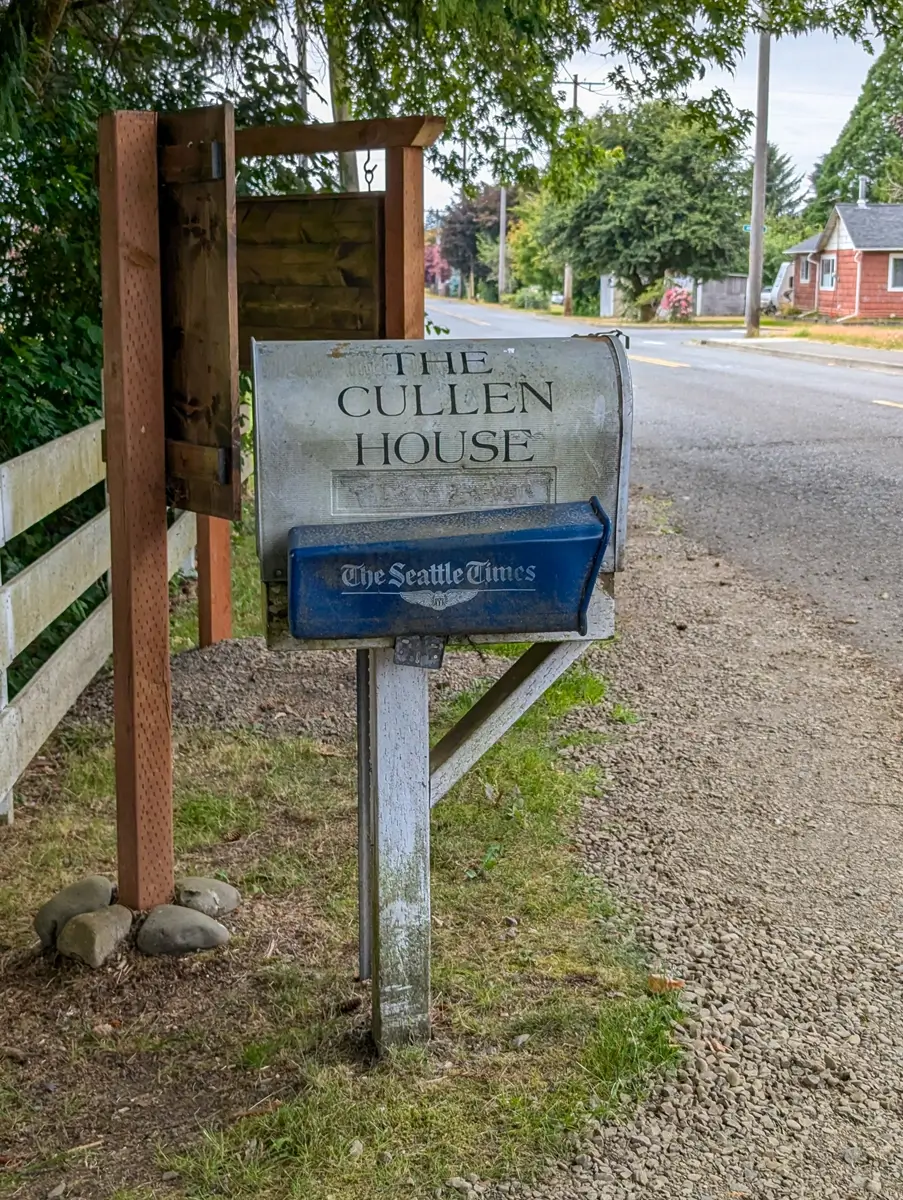 The Cullen House Mailbox A close-up image of a weathered mailbox on a wooden post. The side of the mailbox has the words "THE CULLEN HOUSE" painted on it, and a blue newspaper box below reads "The Seattle Times."