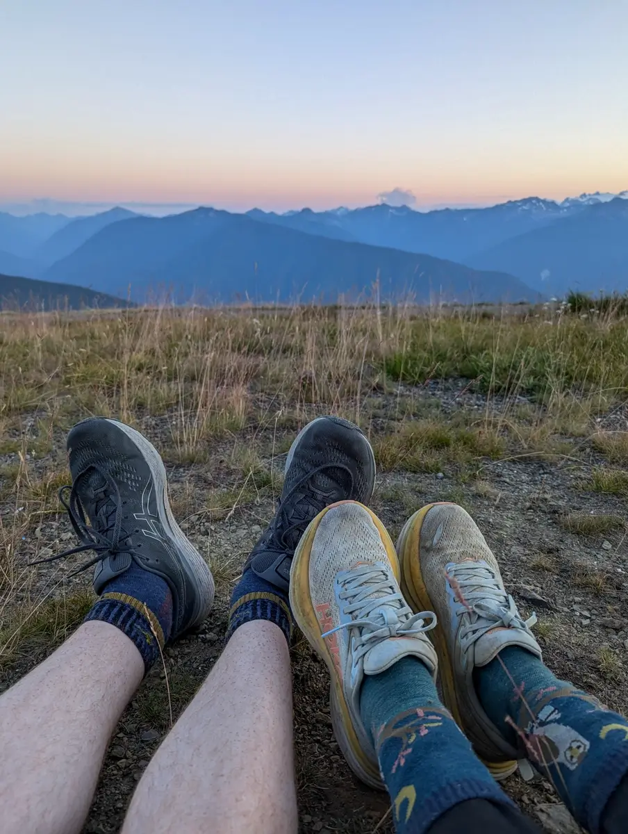 Close-up shot of two pairs of worn hiking shoes and patterned socks resting on the ground at Hurricane Ridge. In the background, the Olympic Mountain Range stretches across the horizon under a soft, twilight sky, symbolizing a completed hike and the reward of the scenic view.