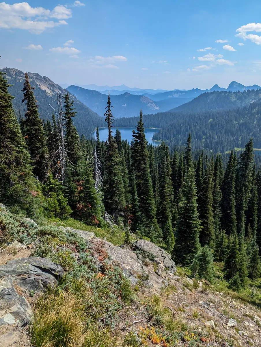 Spectacular vista of Dewey Lake nestled in a valley of dense evergreen forest, viewed from a rocky, high-elevation vantage point on the Pacific Crest Trail (PCT) in Washington. Distant mountain ranges fade into the haze under a clear blue sky, illustrating the expansive scenery of the Mount Rainier National Park area.