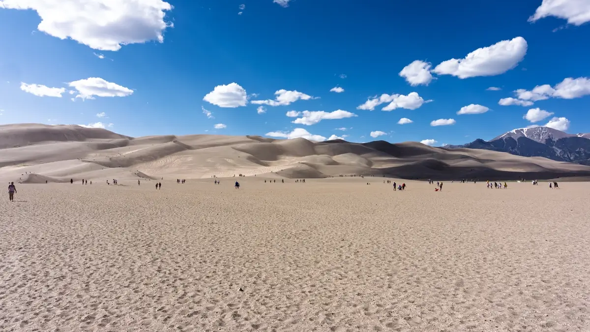 Great Sand Dunes NP From A Distance Panoramic view of Great Sand Dunes National Park, Colorado, showing the massive sand dune field under a bright blue sky with white clouds, with a snow-capped mountain peak visible in the distance. The foreground features a wide, flat, sandy area with small groups of people approaching the base of the colossal dunes, ready for hiking and sandboarding.