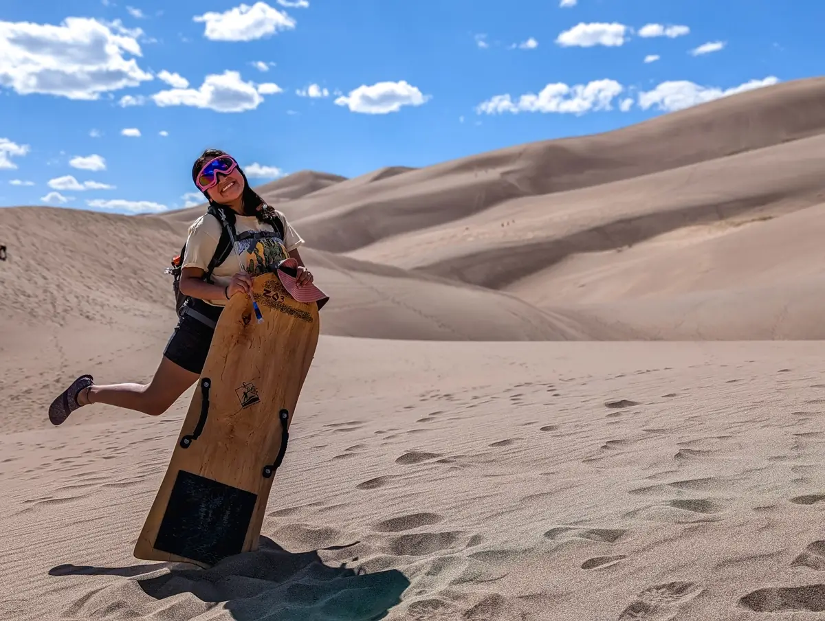 Minerva Posing In Great Sand Dunes Colorado Minerva posing on the enormous sand dunes of Great Sand Dunes National Park, Colorado, wearing goggles and holding a large wooden sandboard, ready for sandboarding Great Sand Dunes National Park. The sun is shining brightly, highlighting the vast, sandy landscape behind her.
