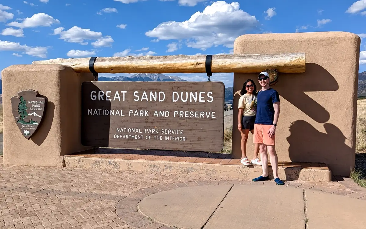 Minerva And Sam At The Colorado Great Sand Dunes National Park Sign Minerva and Sam standing and smiling in front of the rustic, adobe-style entrance sign. Great Sand Dunes National Park and Preserve photos often include this sign, which is made of wood and logs, with mountains and clear skies visible in the background. This is a classic photo opportunity for park visitors.