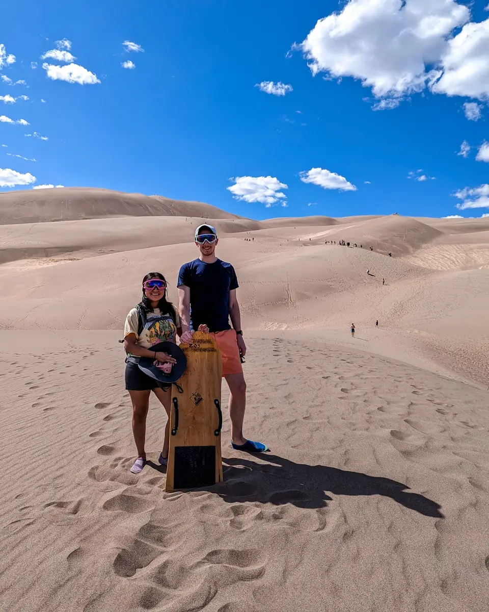 Minerva And Sam In Great Sand Dunes National Park Minerva and Sam standing together on the sandy slopes of Great Sand Dunes National Park, Colorado, holding a wooden sandboard. They are wearing sunglasses and comfortable outdoor gear, with the massive, towering sand dunes and other hikers visible behind them against the blue sky.
