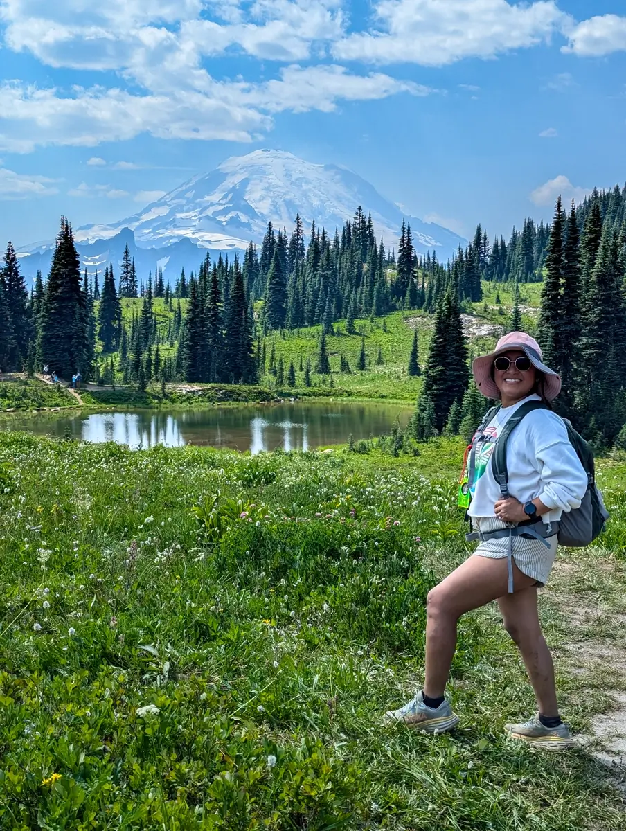 Hiker Minerva stands smiling in the foreground with a backpack and sun hat, posing in front of a majestic view of Mount Rainier and a small alpine lake. The photo highlights the beautiful subalpine meadow and evergreen forest found along the Naches Peak Loop Trail or the Pacific Crest Trail.