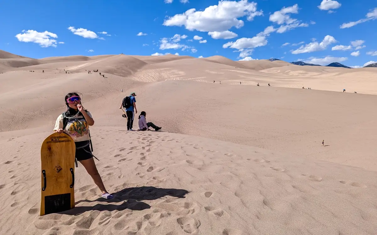 Minerva In Great Sand Dunes Colorado Minerva standing on a sandy slope in Great Sand Dunes National Park, Colorado, holding a sandboard and wearing bright pink goggles under a clear blue, cloudy sky. Footprints cover the foreground, leading up to the massive sand dunes in the background, where other visitors are hiking.