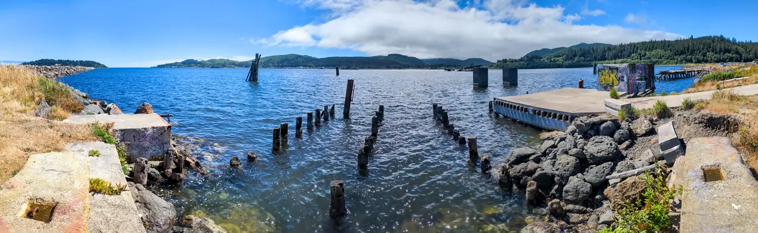 Neah Bay A wide panoramic view of the waterfront at Neah Bay. The bright blue water has several decaying wooden pilings sticking out of it. There is an old, broken concrete dock/platform to the right, and green, forested hills rise in the distance.