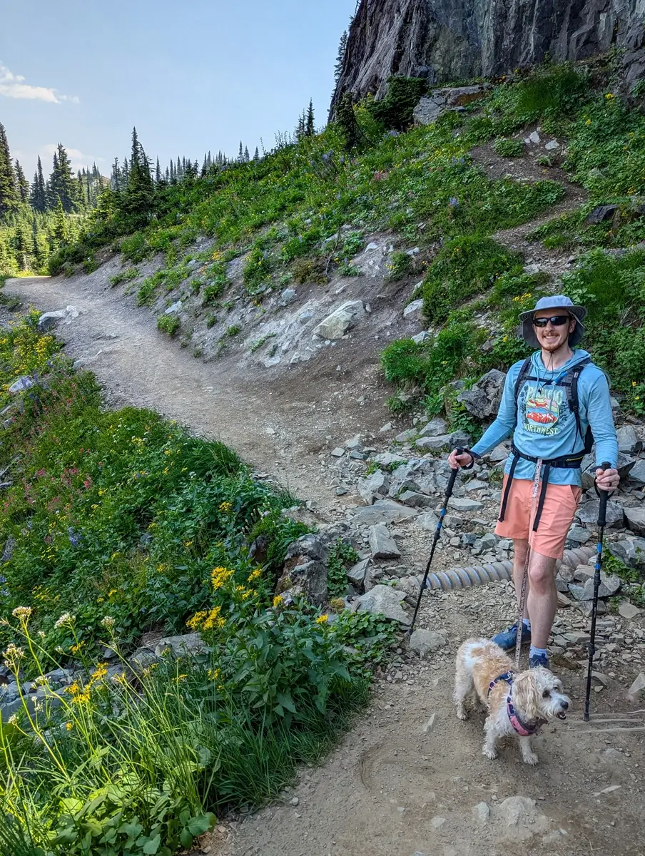 Hiker Sam smiling with his small, shaggy dog Lily on the Pacific Crest Trail in Mount Rainier National Park. They are standing on a rocky trail section, equipped with trekking poles, with a steep, green hillside and cliff face in the background.