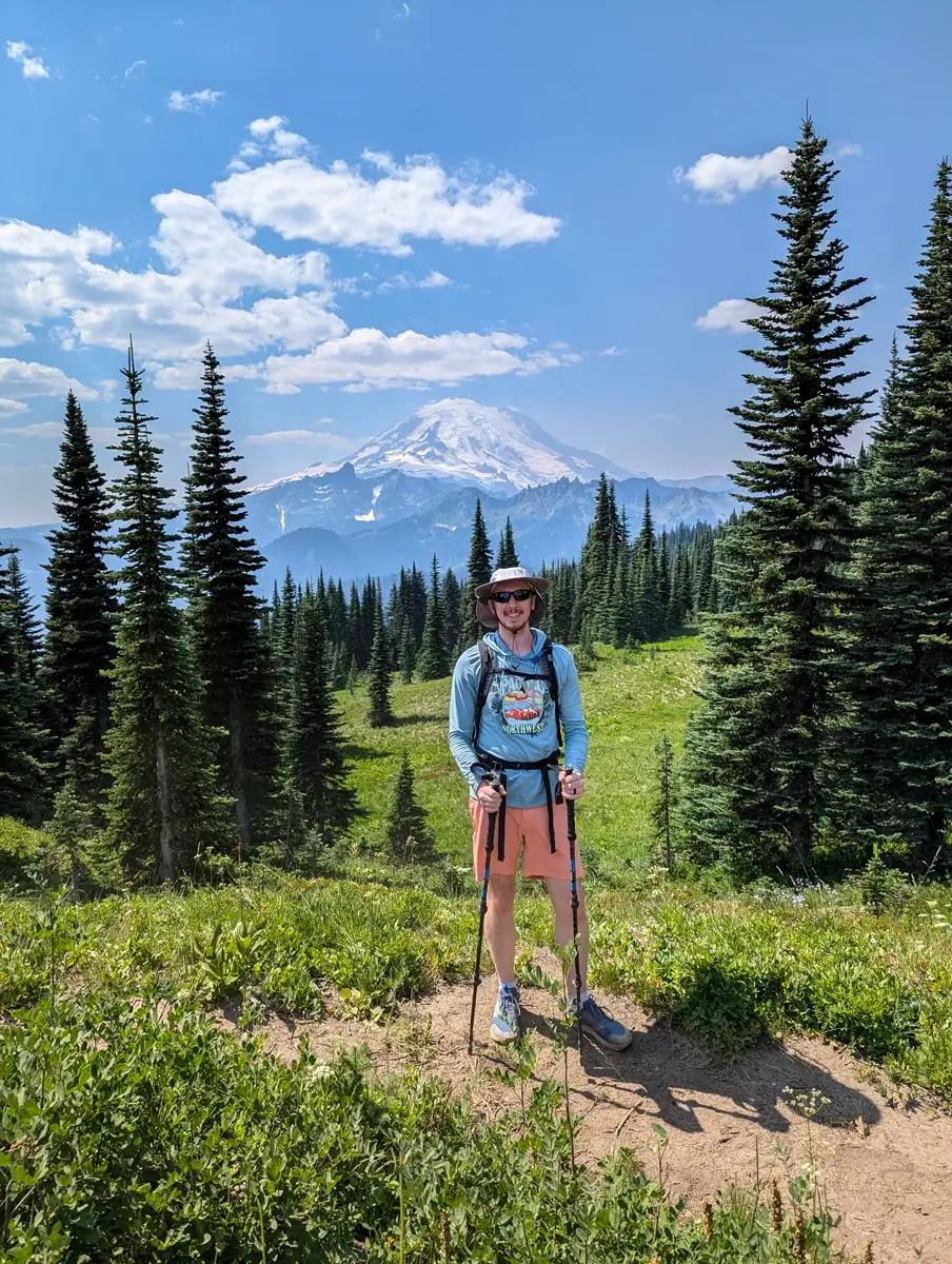 Hiker Sam stands smiling, wearing a hat and holding trekking poles, with the massive, snow-covered Mount Rainier directly behind him. He is on the Naches Peak Loop Trail or Pacific Crest Trail, surrounded by a sunny alpine meadow and tall evergreen trees.