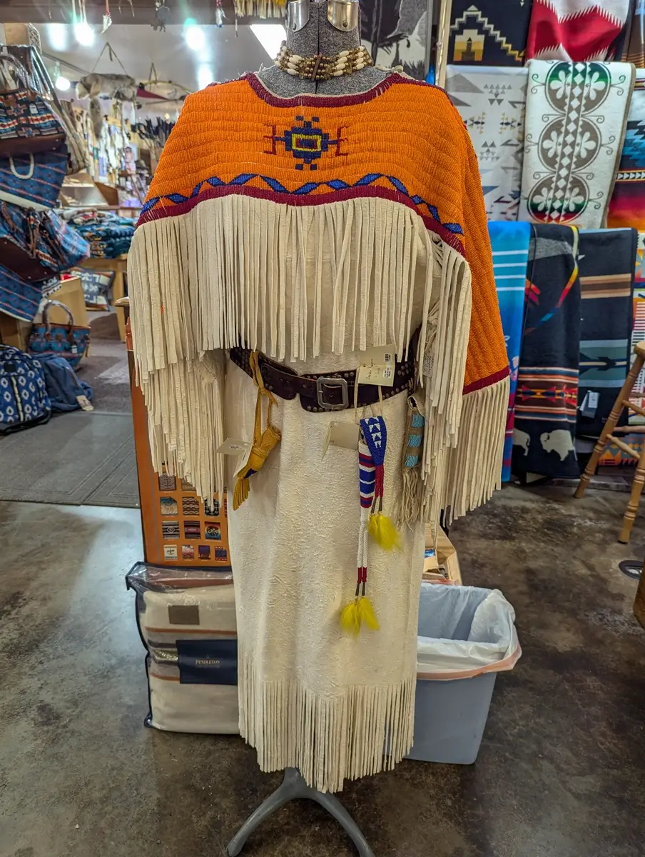 Custer Battlefield Trading Post Cafe Buckskin Dress Mannequin wearing a traditional beaded dress with an orange and blue geometric yoke, long white fringe sleeves, and fringe hem. The dress is styled with a wide belt, beaded accessories, and necklaces inside the trading post surrounded by woven textiles.